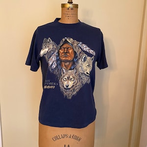 Puede incluir: Una camiseta azul marino con un estampado gráfico de un hombre nativo americano con lobos. El texto "All Others Olfeboro" está impreso en la camiseta.