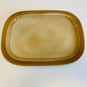 May include: A light brown ceramic serving dish with a raised rim and a textured design. The dish is oval shaped and has a slightly rounded edge.