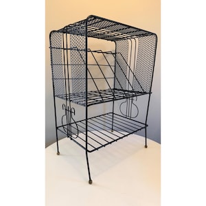 May include: Black metal shelving unit with a two-tiered design. The top tier has a mesh back and a wire grid shelf. The bottom tier has a wire grid shelf. The unit has four legs with brass-colored feet. The unit is designed to hold items.