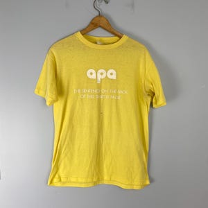 May include: Bright yellow t-shirt with the word "apa" in white block letters. Below the word is the text: "THE SENTENCE ON THE BACK OF THIS SHIRT IS FALSE". The shirt is hanging on a wooden hanger.
