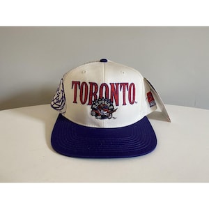 Puede incluir: Gorra de béisbol blanca y morada con la palabra "TORONTO" en rojo y el logotipo de los Toronto Raptors.