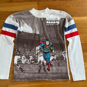 Könnte beinhalten: Weißes Langarmshirt mit einem Vintage-Superman-Druck. Das Shirt hat ein rot-weiß-blaues Streifenmuster an den Ärmeln und den Text "Copyright National Periodical Publications, Inc. 1978" auf der Vorderseite.