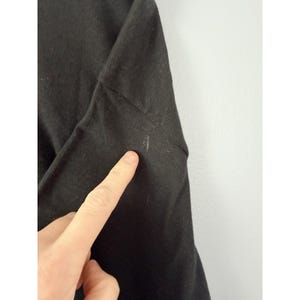 Może przedstawiać: Zbliżenie czarnej koszulki z długim rękawem. Materiał jest ciemny i wydaje się być dzianiną. Palec wskazuje na rękaw, podkreślając małą plamę. Koszulka jest na białym tle.