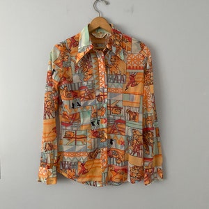 Puede incluir: Una camisa de manga larga con botones y un estampado colorido de tigres y artistas de circo. La camisa está hecha de una tela transparente y tiene un corte holgado.