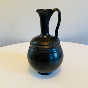 May include: A black ceramic pitcher with a rounded body and a single handle. The pitcher has a narrow neck and a small base.