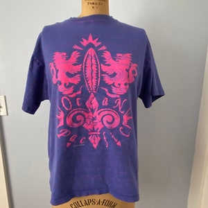 May include: A purple t-shirt with a pink graphic design of a crest with two lions and the words "Ocean Pacific".