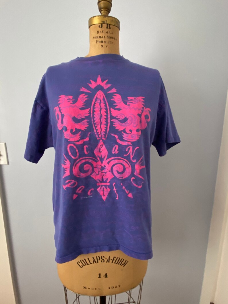 May include: A purple t-shirt with a pink graphic design of a crest with two lions and the words "Ocean Pacific".