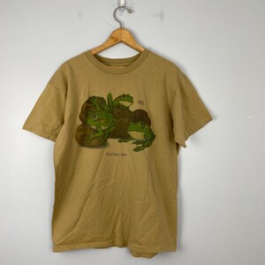 May include: A tan t-shirt with a graphic of four green frogs on a pile of rocks. The text "Frog Haven, Idaho" is printed below the frogs.