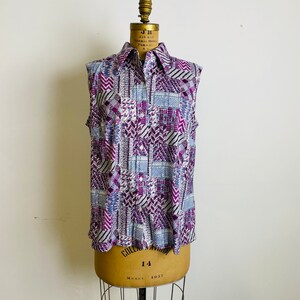 May include: A sleeveless button-up shirt with a geometric pattern in shades of purple, blue, and white. The shirt has a collared neckline and a button-up front.