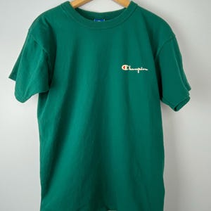 Könnte beinhalten: Ein grünes T-Shirt mit kurzen Ärmeln und einem weißen "Champion"-Logo auf der Brust.