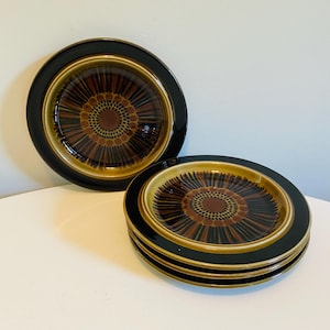 May include: A set of four ceramic plates with a black rim and a brown and gold floral design. The plates have a circular shape and are stacked on top of each other. The plates are on a white surface.