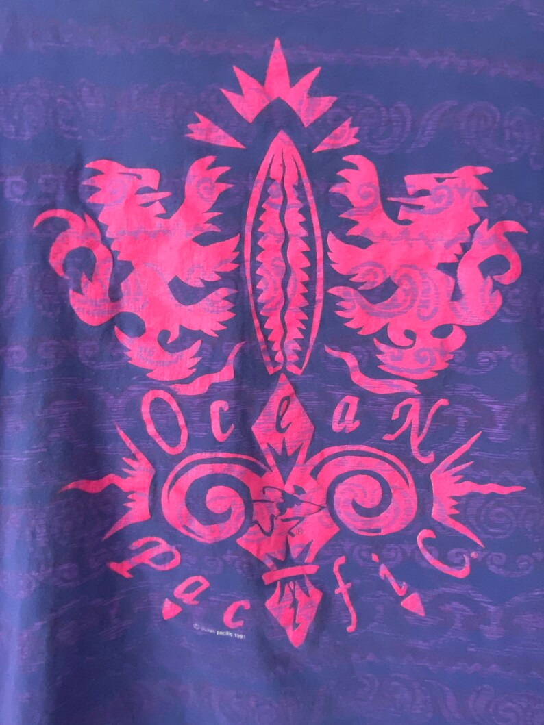 May include: A purple t-shirt with a pink graphic design of a stylized fleur-de-lis with two lions on either side. The graphic says "Ocean Pacific" in a stylized font.