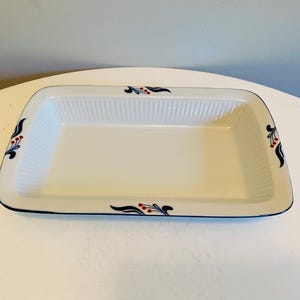 May include: A rectangular, off-white ceramic baking dish with a ribbed interior. The dish has a black rim and is decorated with a stylized floral design in black, red, and blue at each corner. The dish is empty.