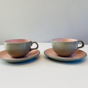 May include: Two gray and pink teacups and saucers. The cups have a light gray band around the middle and a pink interior. The saucers are a light pink with a gray rim.