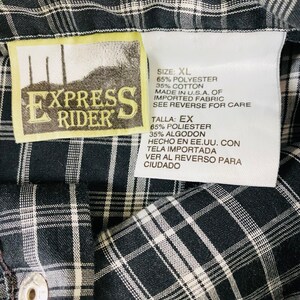 May include: A white tag with black text on a black and white plaid shirt. The tag says "Express Rider" and lists the size as XL. The fabric is 65% polyester and 35% cotton. It was made in the USA of imported fabric. Care instructions are on the reverse side.