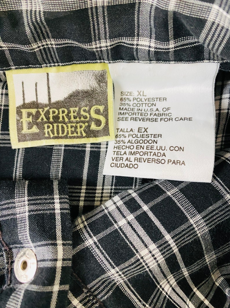 May include: A white tag with black text on a black and white plaid shirt. The tag says "Express Rider" and lists the size as XL. The fabric is 65% polyester and 35% cotton. It was made in the USA of imported fabric. Care instructions are on the reverse side.
