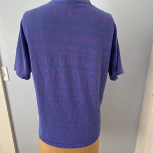 May include: A purple t-shirt with a faded purple graphic print all over. The graphic print is a repeating pattern of text and designs.