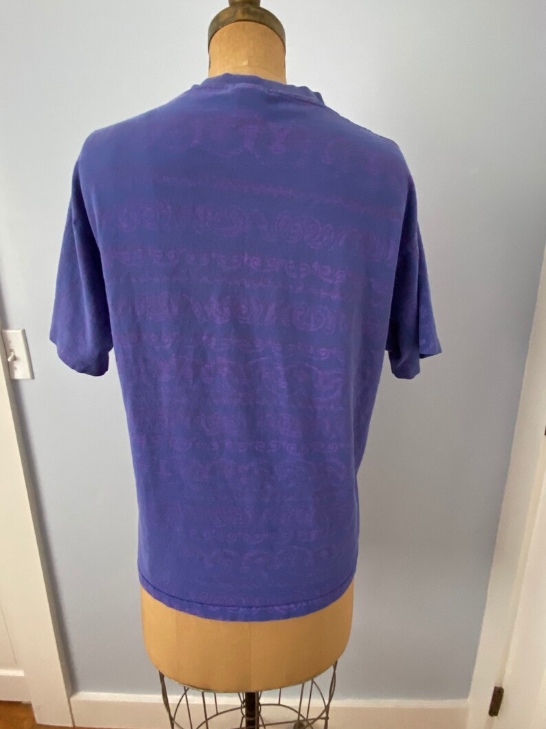 May include: A purple t-shirt with a faded purple graphic print all over. The graphic print is a repeating pattern of text and designs.