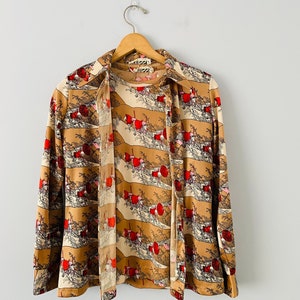 May include: A vintage button-down shirt with a unique landscape print in shades of tan, cream, and red. The long-sleeved shirt is hanging on a wooden hanger. The brand name "Vizzi" is visible on the collar.