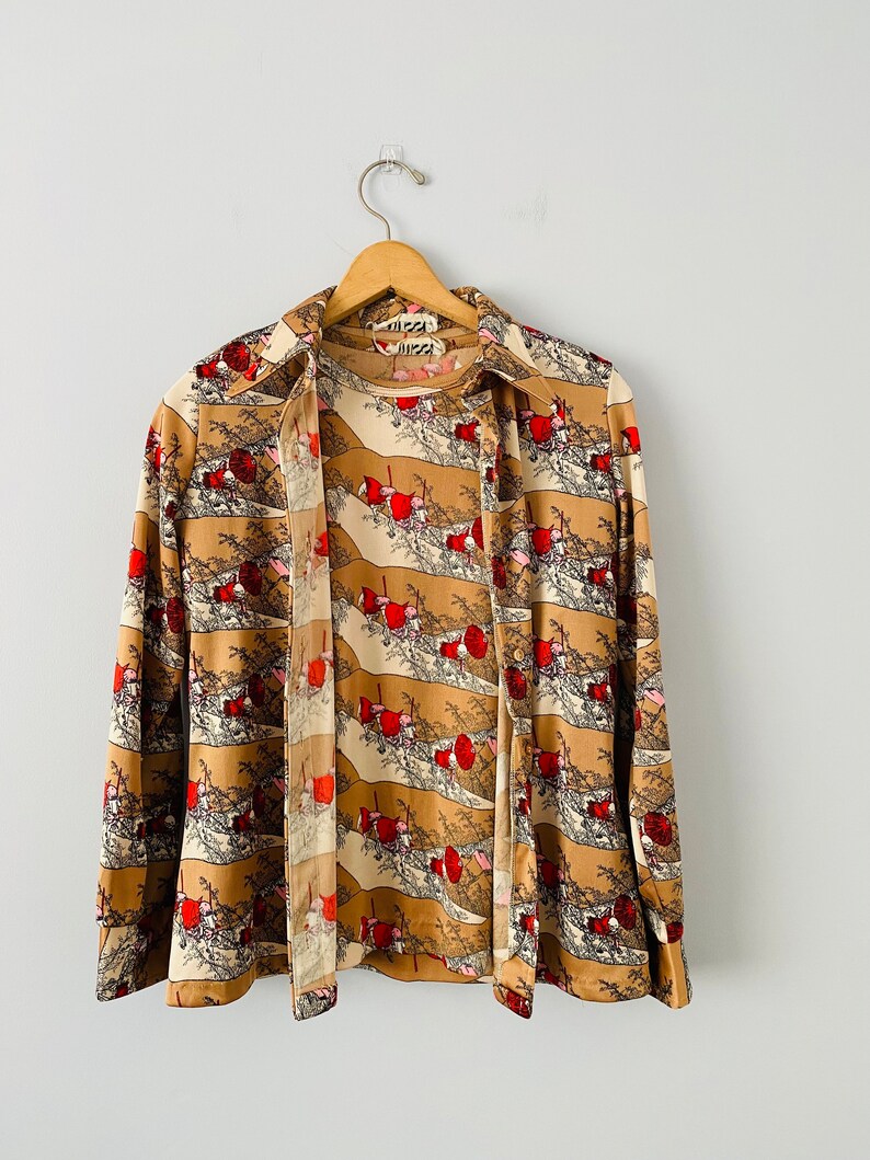 May include: A vintage button-down shirt with a unique landscape print in shades of tan, cream, and red. The long-sleeved shirt is hanging on a wooden hanger. The brand name "Vizzi" is visible on the collar.