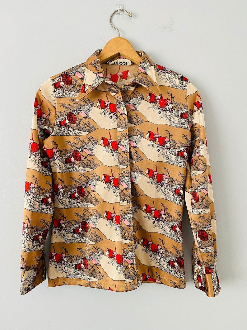 May include: A vintage button-down shirt with a repeating landscape pattern in tan, cream, red, and black. The long-sleeved shirt is hanging on a wooden hanger. The shirt has a collared neckline and buttoned cuffs.