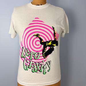 Peut inclure: T-shirt blanc avec un graphisme d'un skateur en l'air, sur fond de spirale rose et blanche. Les mots "Zero Gravity" sont en vert et soulignés de noir. Le t-shirt est présenté sur un mannequin.