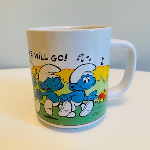 Op de afbeelding: Witte keramische mok met een illustratie van twee Smurfen die elkaars hand vasthouden, met muzieknoten en de tekst "WE WILL GO!". De mok heeft een wit handvat en een groen, geel en blauw landschapsontwerp.