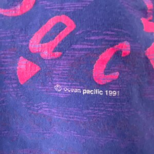 May include: Close-up of a purple and pink t-shirt with the word "Back" written in pink. The shirt has a textured fabric and the text "Ocean Pacific 1991" is visible at the bottom.