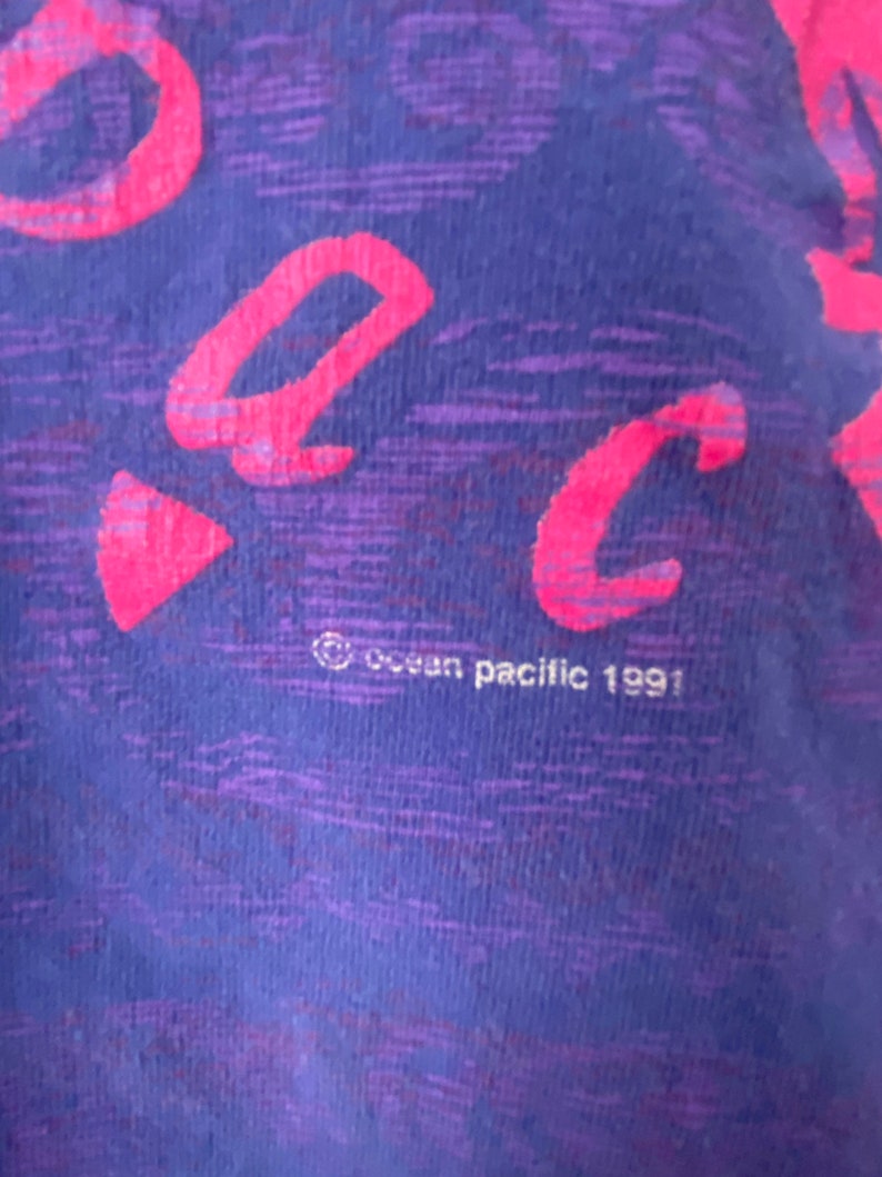 May include: Close-up of a purple and pink t-shirt with the word "Back" written in pink. The shirt has a textured fabric and the text "Ocean Pacific 1991" is visible at the bottom.