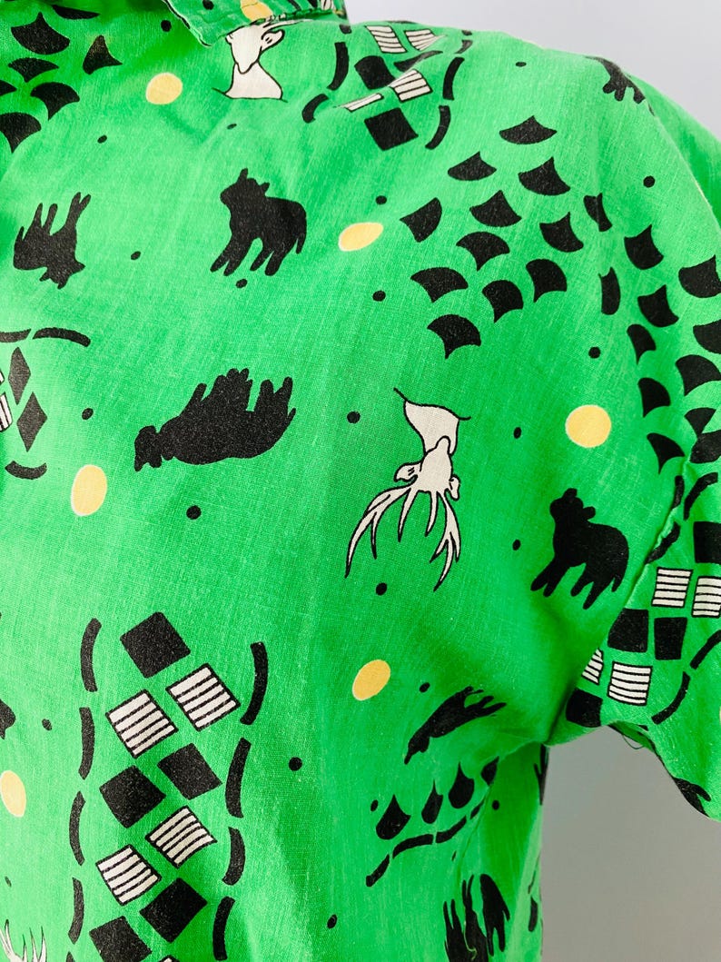 Puede incluir: Camisa verde brillante de manga corta con un patr&oacute;n repetido de siluetas de animales negros, formas geom&eacute;tricas y puntos amarillos. El dise&ntilde;o incluye ciervos, osos y formas abstractas. La tela parece ser ligera.