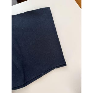 Może przedstawiać: Zbliżenie fragmentu granatowej tkaniny, prawdopodobnie części odzieży. Tkanina jest gładka i wydaje się być w jednolitym kolorze. Widoczny jest brzeg tkaniny, pokazujący złożony d&oacute;ł.