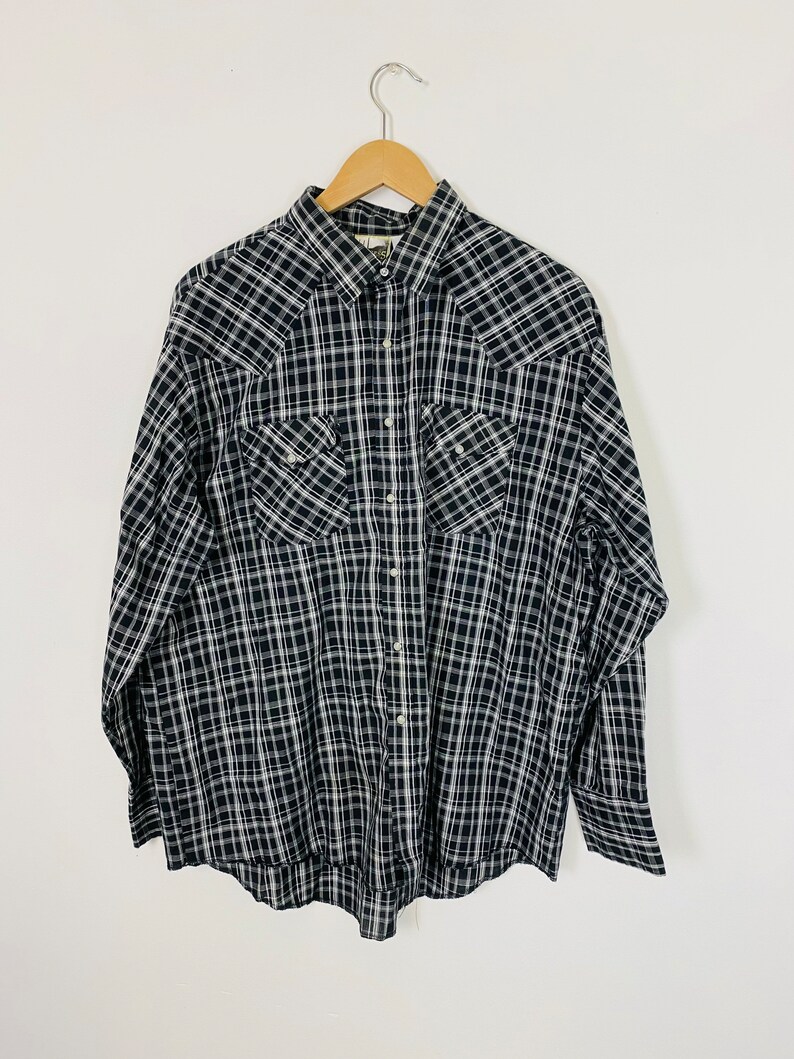 May include: A black and white plaid long-sleeved button-up shirt with two chest pockets. The shirt is hanging on a wooden hanger.