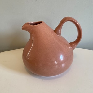 May include: A pink ceramic pitcher with a rounded body and a curved handle. The pitcher has a smooth, glossy finish.