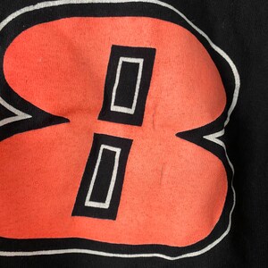 May include: A black t-shirt with a large, white-outlined, orange number "8" printed on the front. The number is stylised with two white rectangles inside the orange shape.