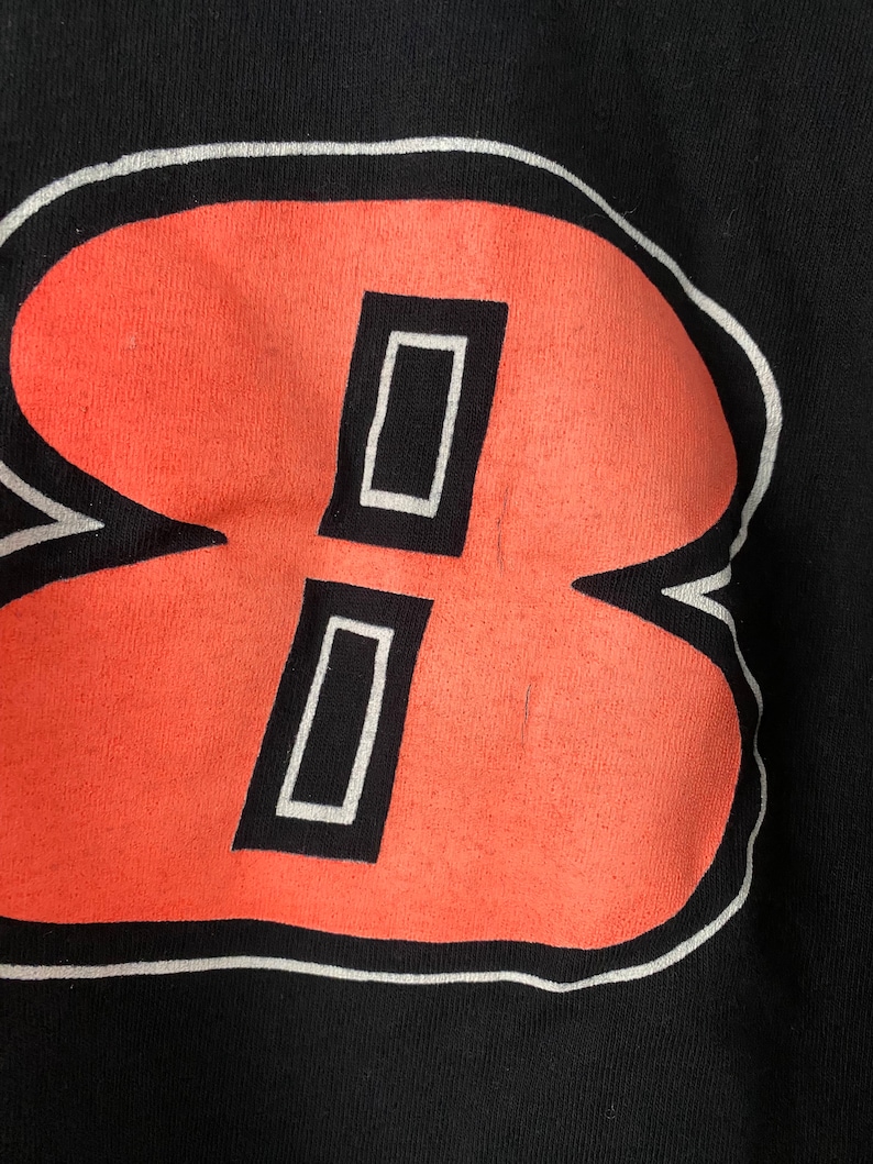 May include: A black t-shirt with a large, white-outlined, orange number "8" printed on the front. The number is stylised with two white rectangles inside the orange shape.