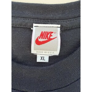 Może przedstawiać: Zbliżenie kołnierza czarnej koszulki z metką Nike. Etykieta zawiera logo Nike w kolorze czerwonym i napis "NIKE" w kolorze białym. Poniżej napis "MADE IN U.S.A." i "XL".