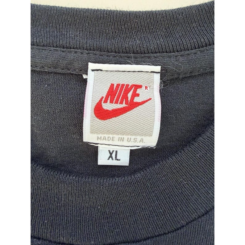 Może przedstawiać: Zbliżenie kołnierza czarnej koszulki z metką Nike. Etykieta zawiera logo Nike w kolorze czerwonym i napis "NIKE" w kolorze białym. Poniżej napis "MADE IN U.S.A." i "XL".