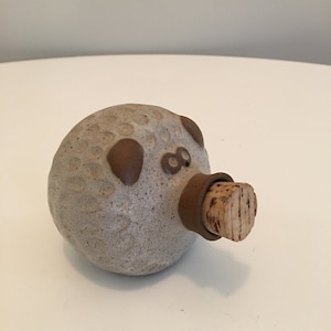 Puede incluir: Una pequeña botella de cerámica con un tapón de corcho. La botella tiene forma de cerdo y tiene una superficie texturizada. El cerdo tiene ojos marrones y una nariz marrón.