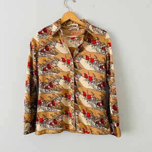 May include: A button-down shirt with a repeating pattern of red, white, and tan landscape scenes. The long-sleeved shirt is hanging on a wooden hanger against a white background. The collar and cuffs are a solid tan color.