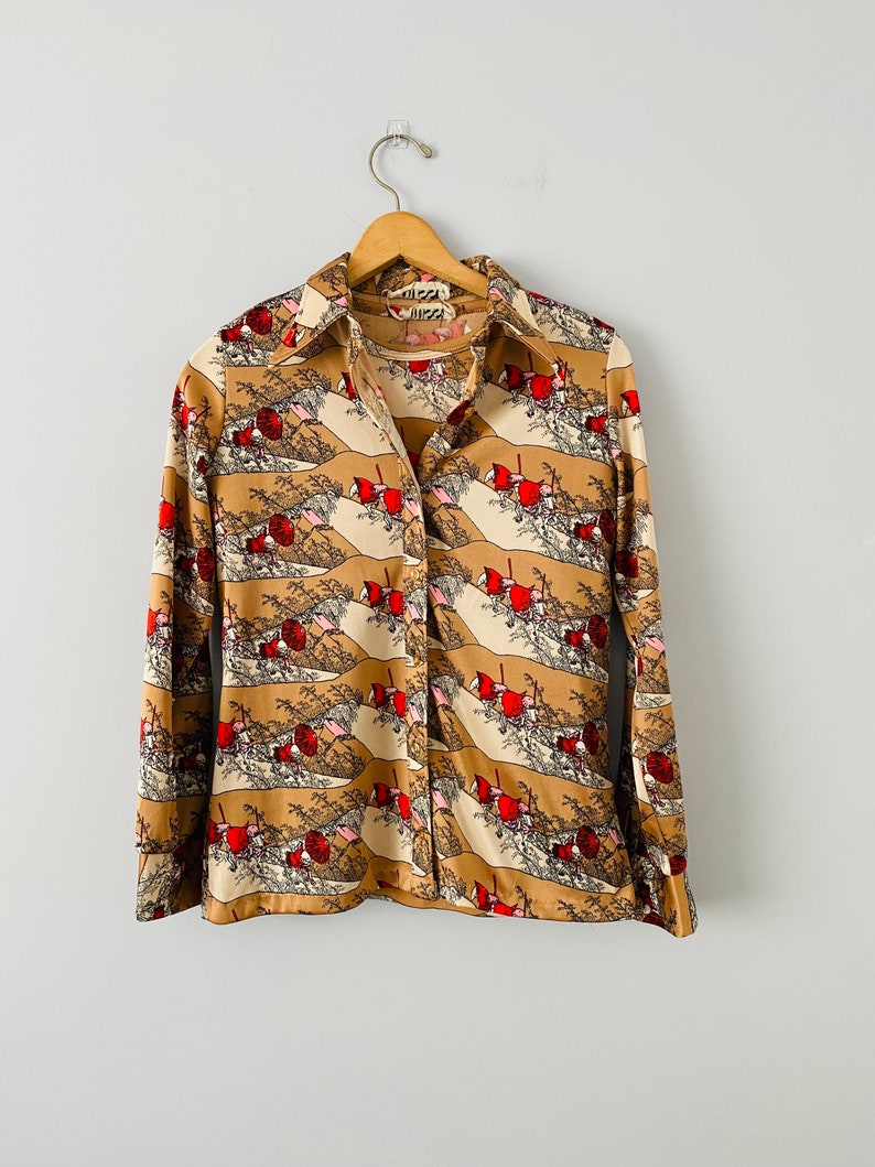 May include: A button-down shirt with a repeating pattern of red, white, and tan landscape scenes. The long-sleeved shirt is hanging on a wooden hanger against a white background. The collar and cuffs are a solid tan color.