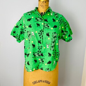 Puede incluir: Una camisa verde de manga corta con un estampado repetido de siluetas de osos y ciervos negros, diamantes y puntos amarillos. La camisa se exhibe en un maniqu&iacute; vintage.