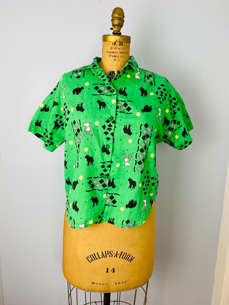 Puede incluir: Una camisa verde de manga corta con un estampado repetido de siluetas de osos y ciervos negros, diamantes y puntos amarillos. La camisa se exhibe en un maniqu&iacute; vintage.