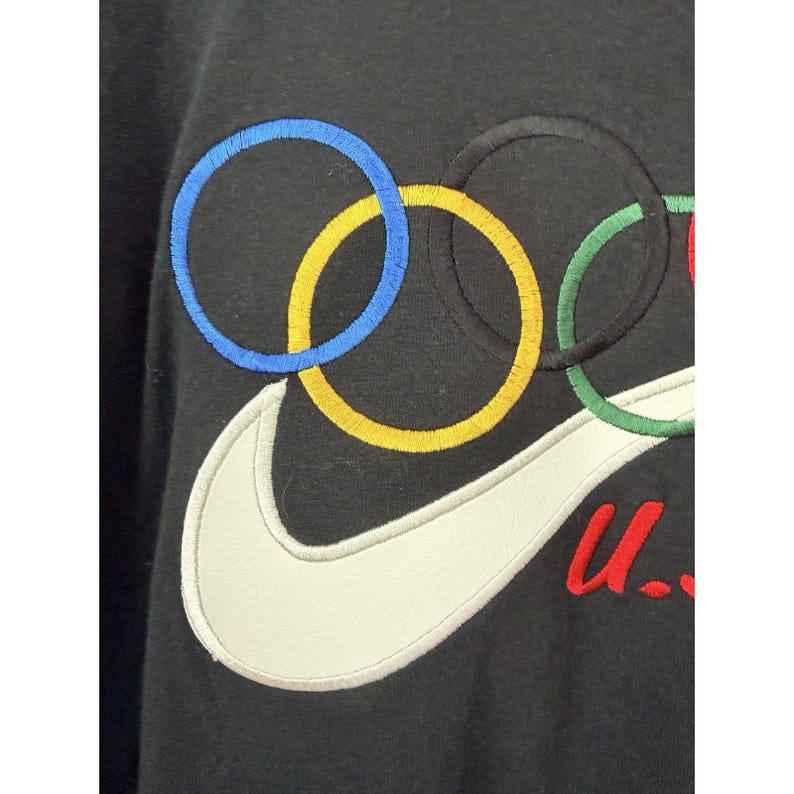Może przedstawiać: Czarna bluza z białym logo Nike i olimpijskimi kołami w kolorze niebieskim, ż&oacute;łtym, czarnym i zielonym. Litery "u." są wyhaftowane na czerwono. Odzież sportowa.