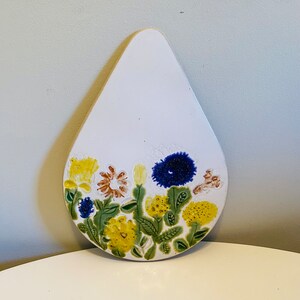Może przedstawiać: Biały ceramiczny podkładka w kształcie łzy z kwiatowym wzorem. Wzór przedstawia żółte, niebieskie i brązowe kwiaty z zielonymi liśćmi.