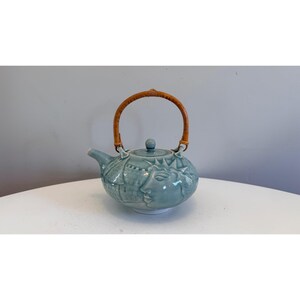 May include: A light blue ceramic teapot with a decorative face design and a woven handle. The teapot has a spout and a lid with a small knob. The handle is made of a light brown woven material.
