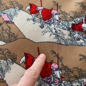 May include: Close-up of a fabric with a repeating pattern of figures on horseback, wearing red cloaks and pink hats. The background features stylized brown and beige hills with sparse trees. The fabric has a textured appearance.