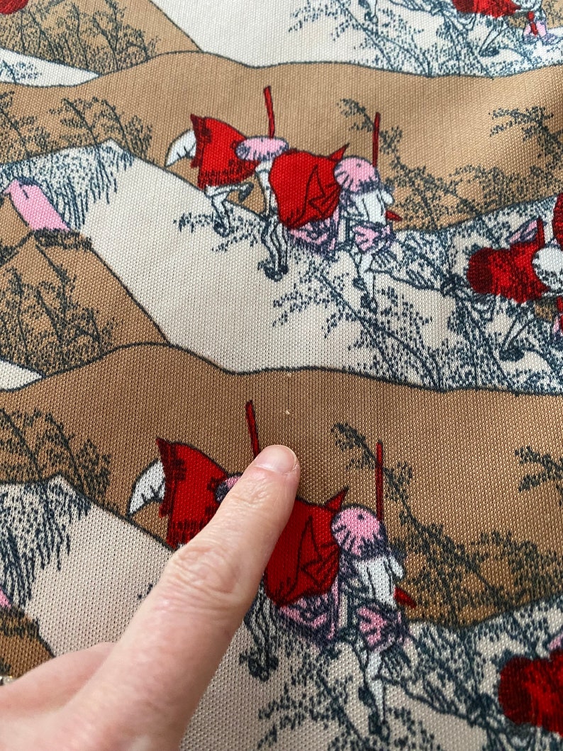 May include: Close-up of a fabric with a repeating pattern of figures on horseback, wearing red cloaks and pink hats. The background features stylized brown and beige hills with sparse trees. The fabric has a textured appearance.