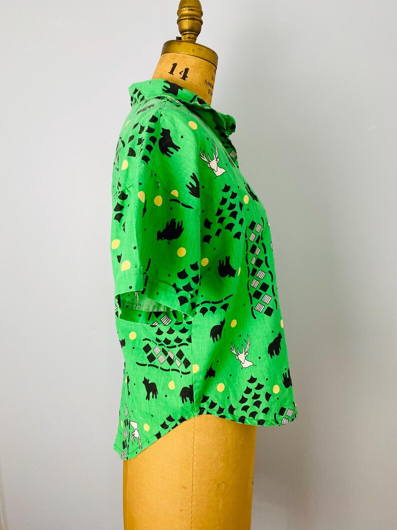 Puede incluir: Una camisa verde brillante con un estampado caprichoso de animales negros, formas geom&eacute;tricas y lunares amarillos. La camisa de manga corta se exhibe en un maniqu&iacute;.