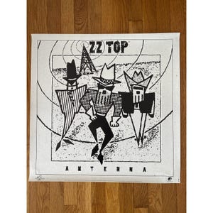 May include: Black and white poster featuring the band ZZ Top. The poster has a graphic illustration of three band members in hats and sunglasses. The word "ANTENNA" is printed at the bottom of the poster.
