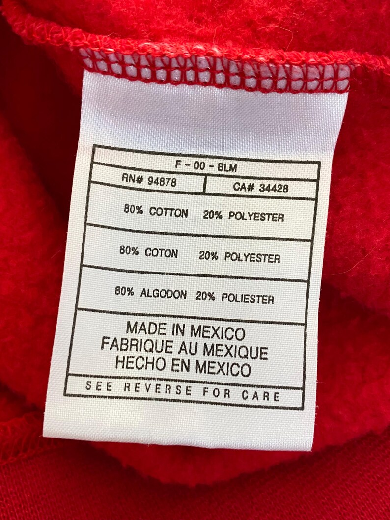 May include: White tag with black text detailing the fabric composition of a red sweatshirt. The tag reads "F-00-BLM", "RN# 94878", "CA# 34428", "80% COTTON 20% POLYESTER", "80% COTON 20% POLYESTER", "80% ALGODON 20% POLIESTER", "MADE IN MEXICO", "FABRIQUE AU MEXIQUE", "HECHO EN MEXICO", and "SEE REVERSE FOR CARE".